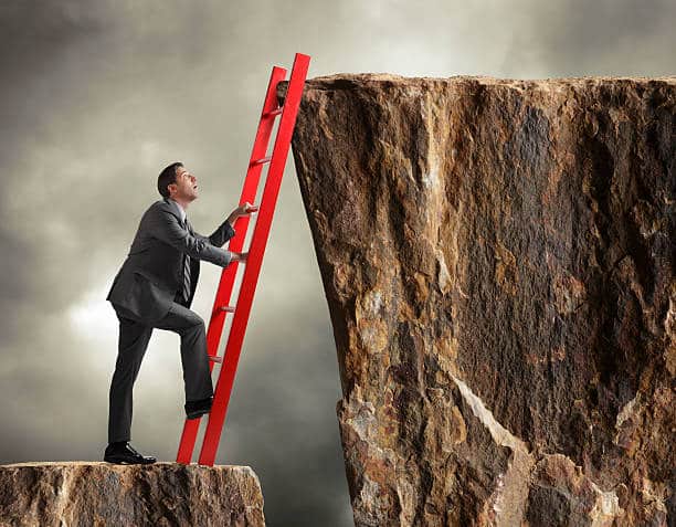 A businessman leans a red ladder against a cliff as he is about to use it in order climb up to another cliff that has a higher vantage point. The businessman has one foot on the first rung of the ladder as he looks up towards his destination. Ominous dark gray clouds are in the background.
