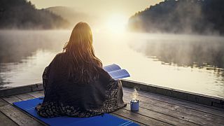 Young woman sitting on a pier and relaxing with book and coffee on sunrise.